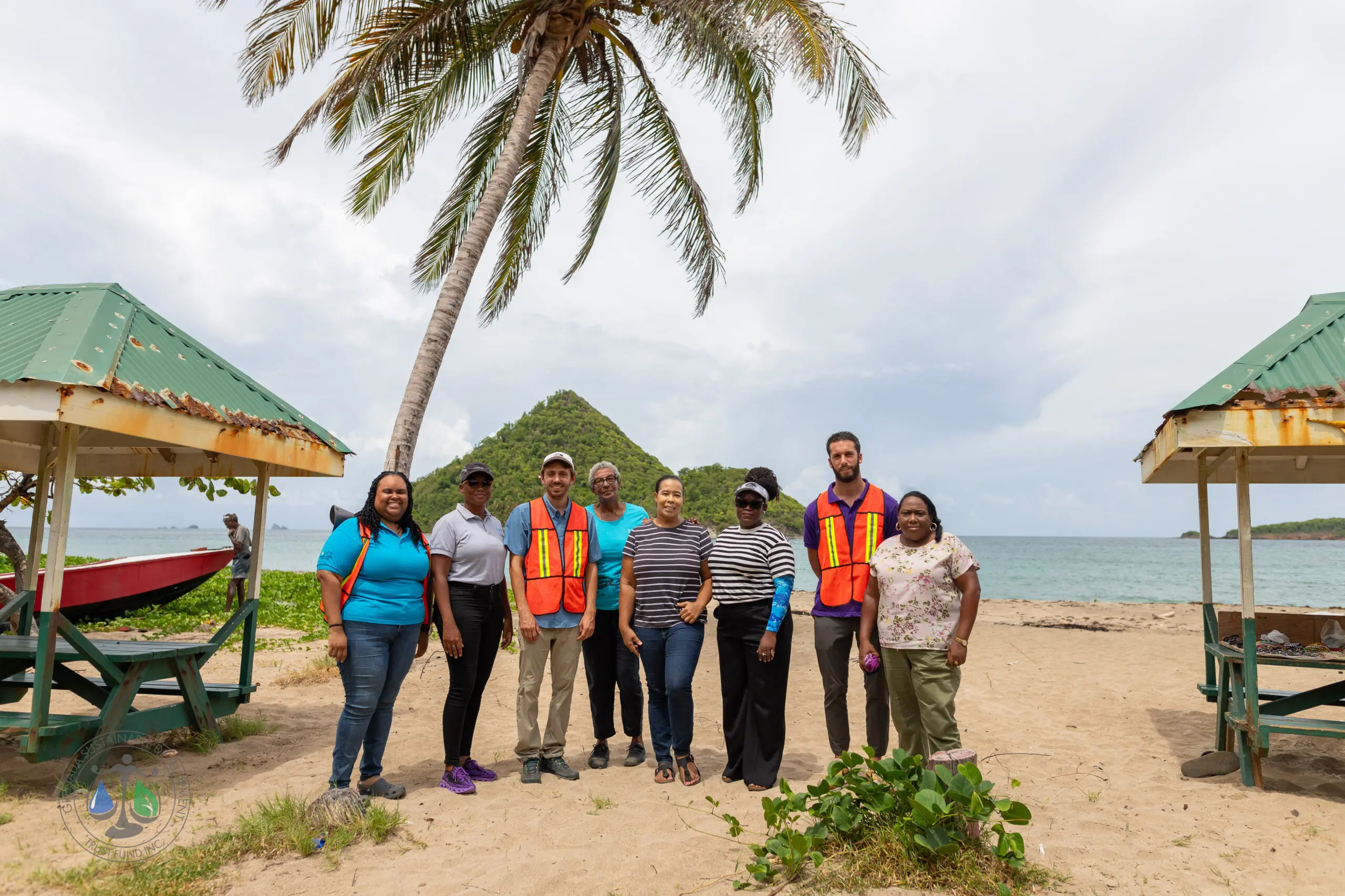 A group of people standing together on Levera Beach in Grenada, with a palm tree and the iconic Sugar Loaf Island in the background. Two individuals are wearing orange safety vests, and the group poses between two green-roofed shelters by the seaside.